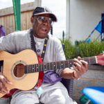 Fernando, a black man wearing sunglasses, playing an acoustic guitar.