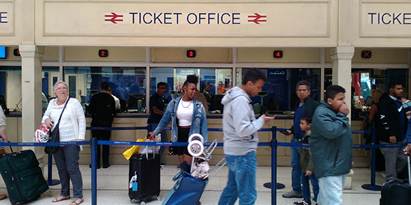 A queue for a busy ticket office, with several people standing in line. There is a sign above the window says Ticket Office with two symbols for British Rail