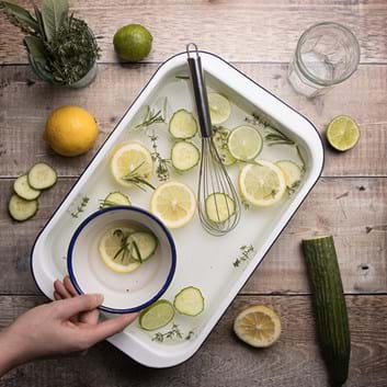 A white tray filled with sliced lemons, limes, cucumbers, ice, and a large balloon whisk