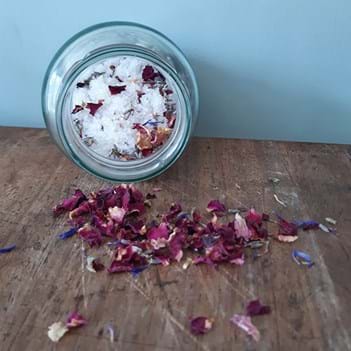 A jar filled with salts and petals on its side, in front of it are many decorative pink petals