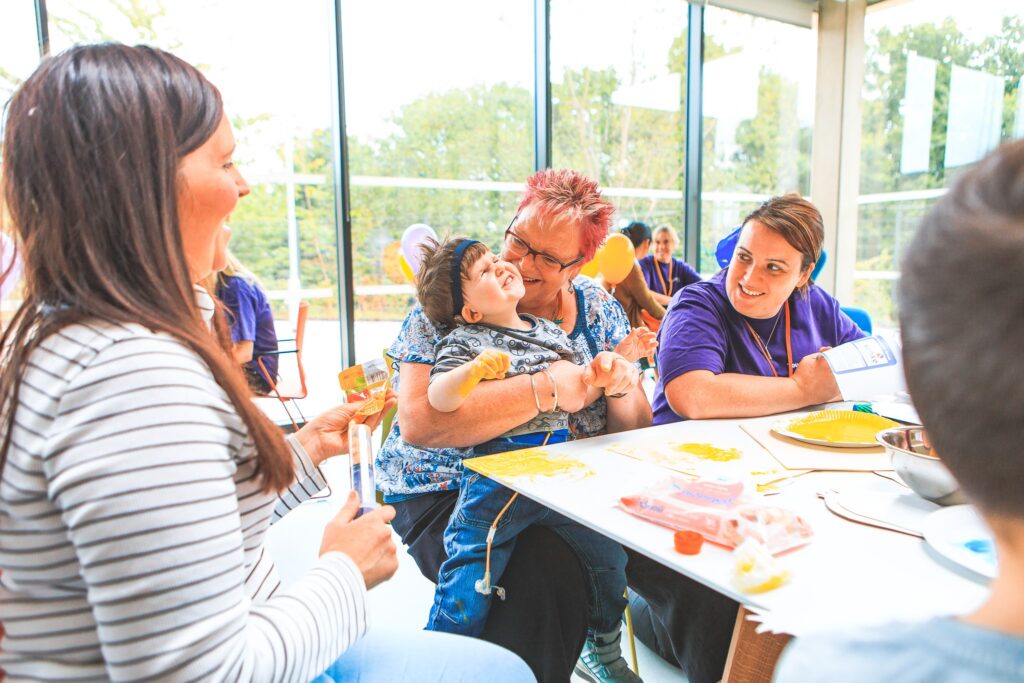 A group of people sit around a table. In the middle, a woman with pink hair is holding a young boy. It's Anne, holding Luca. They're both smiling