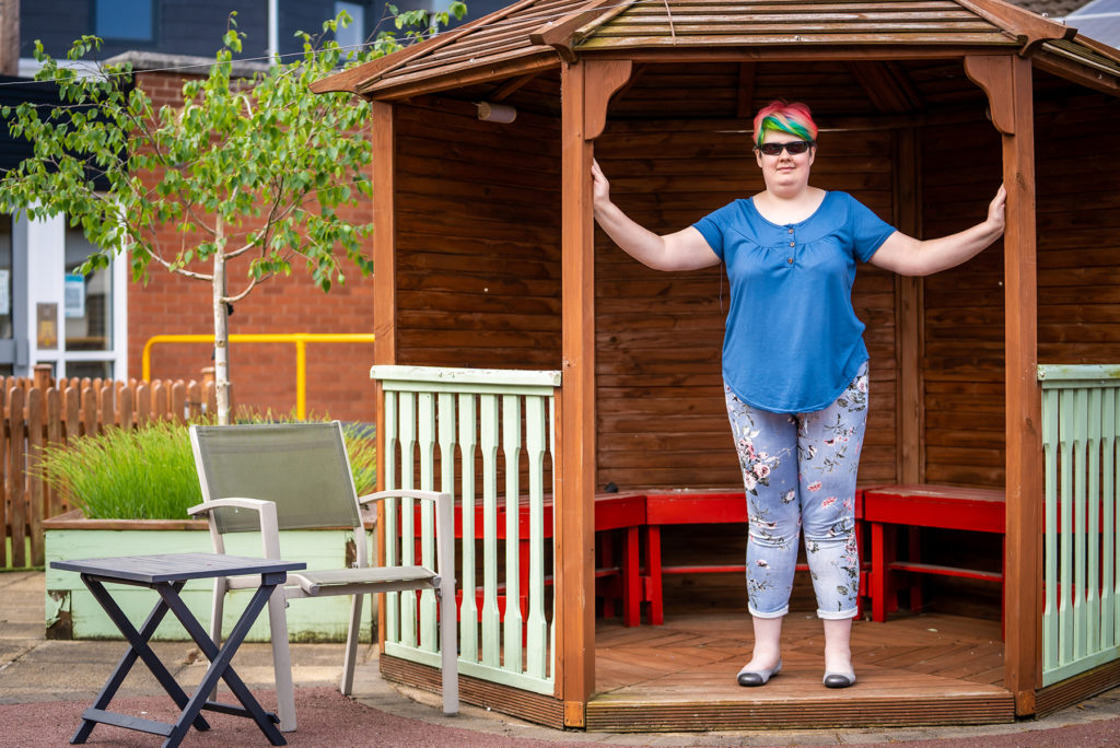A woman is standing on the edge of a hexagonal wooden shelter. She's got her arms out holding two wooden columns. She's wearing leggings, a blue top and has multi-coloured hair