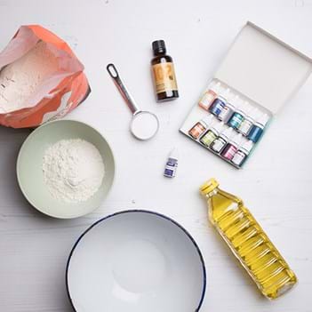 A bottle of yellow oil, two bowls, flour in a red packet, paints, essential oils and a measuring spoon