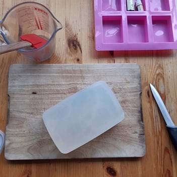 Clear soap on a wooden chopping block, next to a knife, a jug with a spatula in it and a pink soap mould