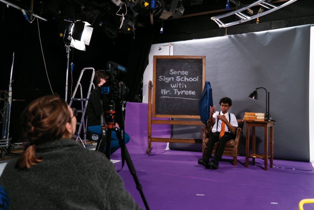 A television studio. We see a woman behind a camera, and a lit area. In the lit area, there's a blackboard with 'Sense Sign School with Mr Tyrese' written in white chalk. Next to the sign, a young man is sitting down wearing a white shirt and an orange tie. It's Mr Tyrese