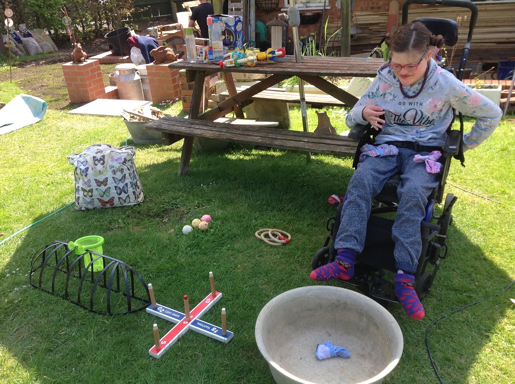 A girl in a wheel chair is wearing a grey jumper outside in the sun. In front of her there is lots of equipment for lots of activities