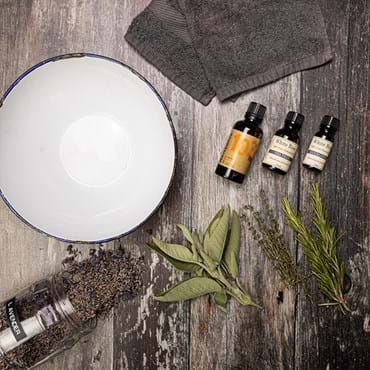 A table top is shown with a bowl, some essential oils and a selection of dried herbs and flowers. / --