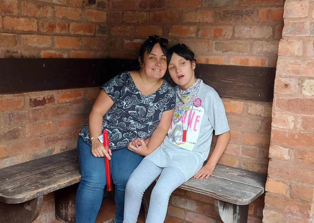 Olivia and her mum, Sam,  holding hands and sitting close together on a corner bench in a brick building. 