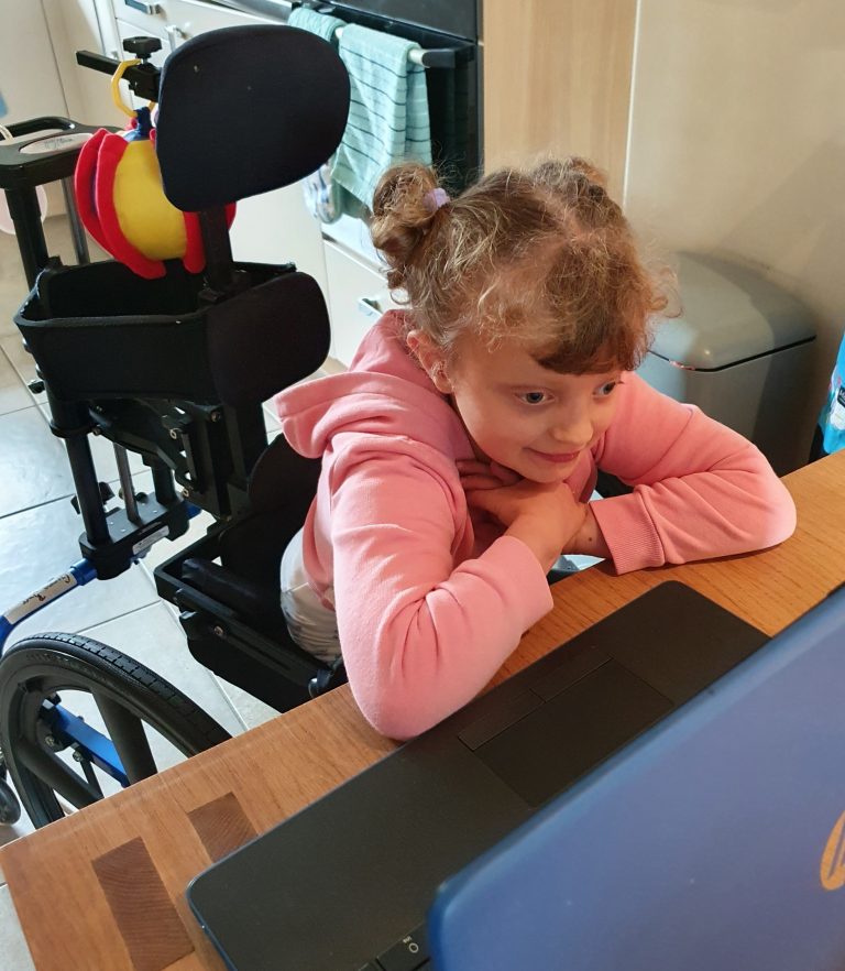 A little girl in a wheelchair sitting at a kitchen table watching a laptop. She has a smile on her face.