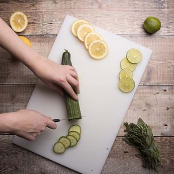 A hand slicing up a cucumber on a chopping board with slices of lemons, and limes