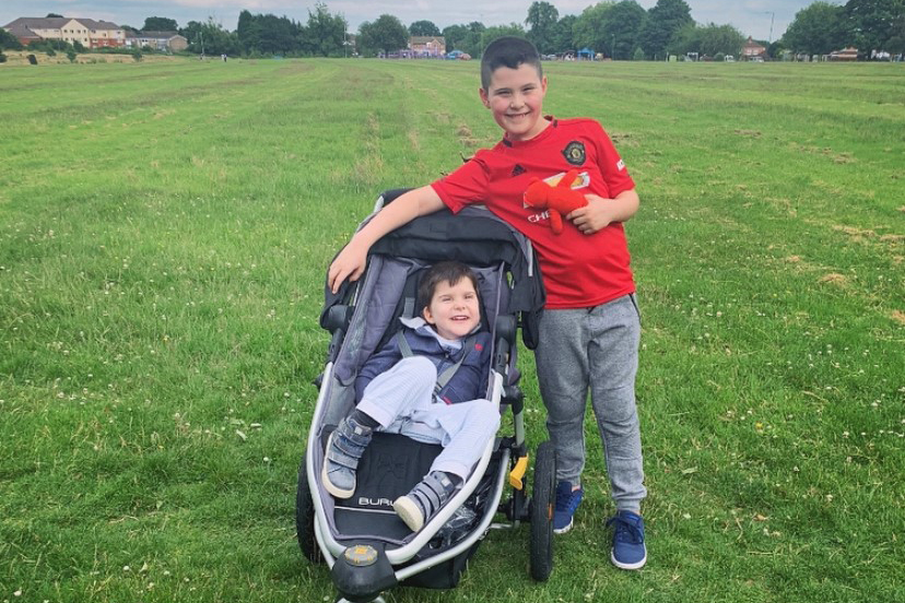 Zach, in a red Manchester United football top, stands next to Luca, who is in a pushchair and wearing a blue top. They are both smiling.