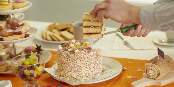 A slice is cut from a cake decorated with sweets and sprinkles, on a table full of more cakes.