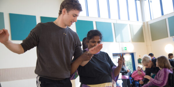 A man is demonstrating a dance pose for the lady stood next to him, she holds his arm as she replicates the move
