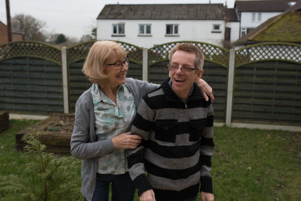 An older woman has her arms around her adult son's shoulders. They're standing in their garden.