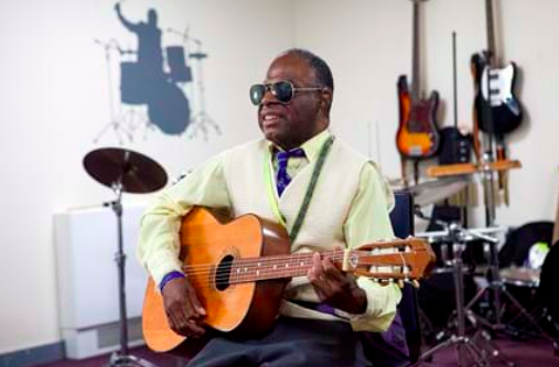 A man with sunglasses on sits in a music room playing guitar. 
