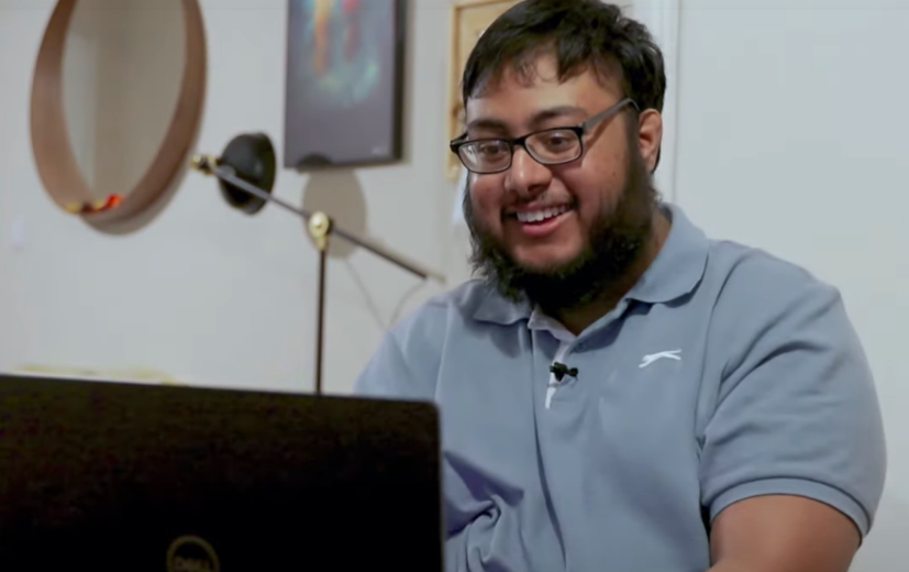 A young man with a beard and glasses smiling as he types on a laptop. 