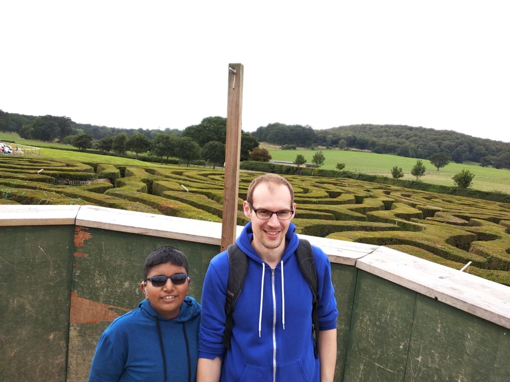 Jack, a Sense holiday volunteer and his buddy are standing in countryside surroundings - in the background is a hedgemaze!