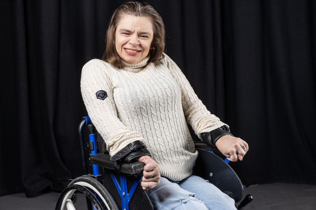 Lisa Simpson, a white woman in a wheelchair with brown hair wearing a white jumper, smiling for the camera.