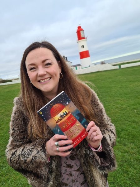 A grinning woman with long brown hair is holding a copy of her book, it has a lighthouse on the cover and there's a real life lighthouse in the background!
