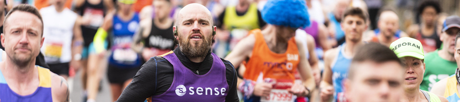 A man with a beard runs in a Sense running vest. There's a sea of people around him.