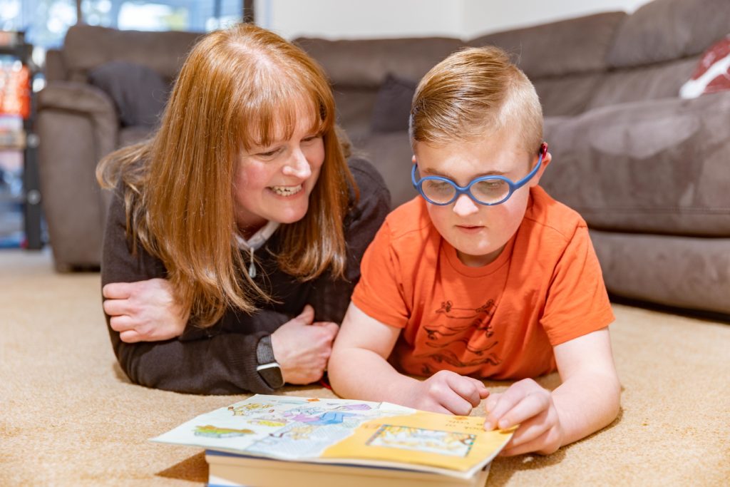A woman and her son lie on the floor, reading a book. The mother, Sarah, is smiling. The boy, Harry, who has blue glasses and an orange t-shirt on, is concentrating on the book.