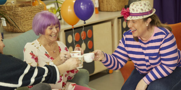 Three women tap their tea cups together while smiling. There's food on a table and balloons in the background.