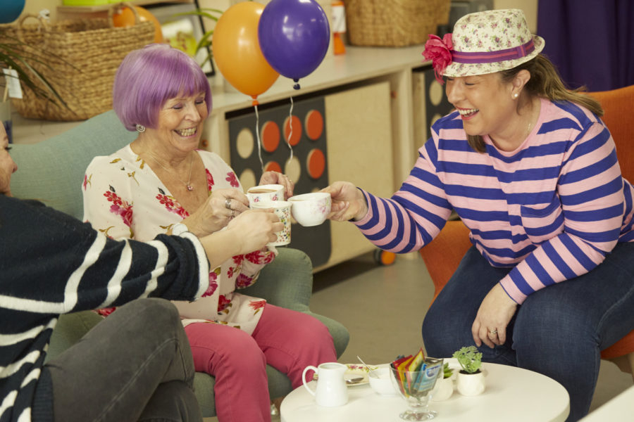 Three women tap their tea cups together while smiling. There's food on a table and balloons in the background.