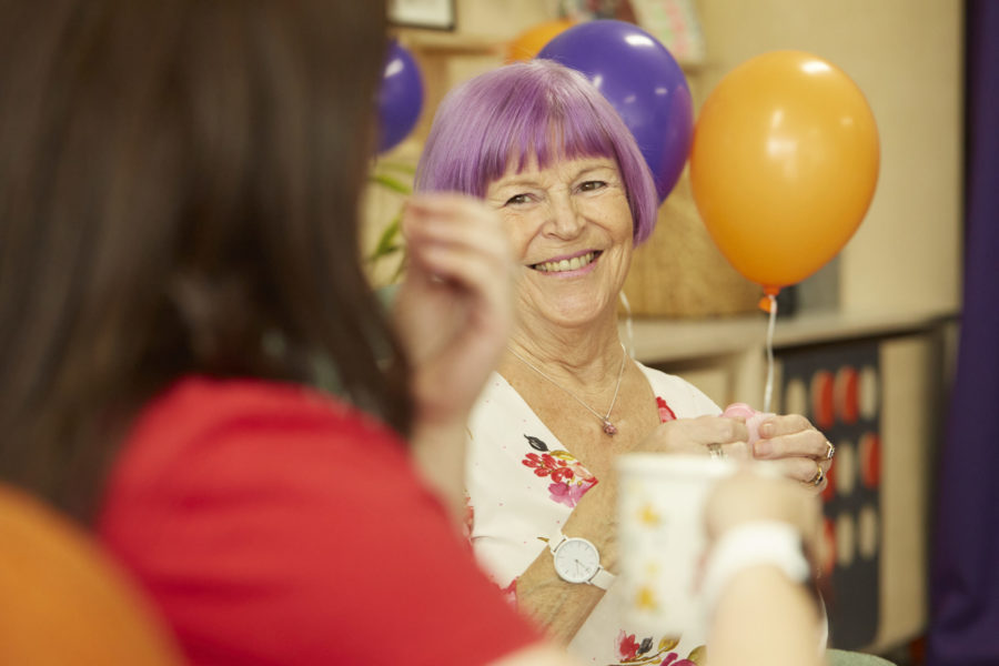 A woman with pink hair smiles as she holds a cup of tea. There are balloons behind her.
