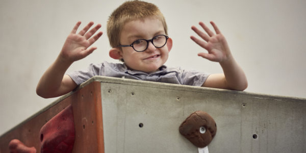 A young boy wearing glasses waves both hands at the camera.