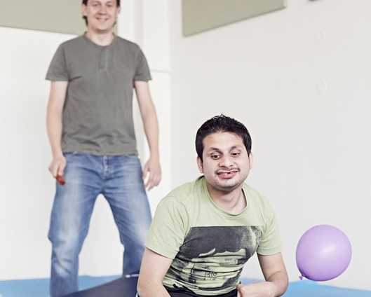 A young man wearing a khaki coloured teeshirt is sitting on the floor smiling at a hovering purple balloon. 