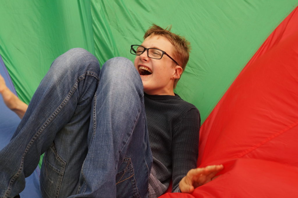 A laughing young boy in glasses and jeans is lying on his back propped up his hands on a green and red parachute