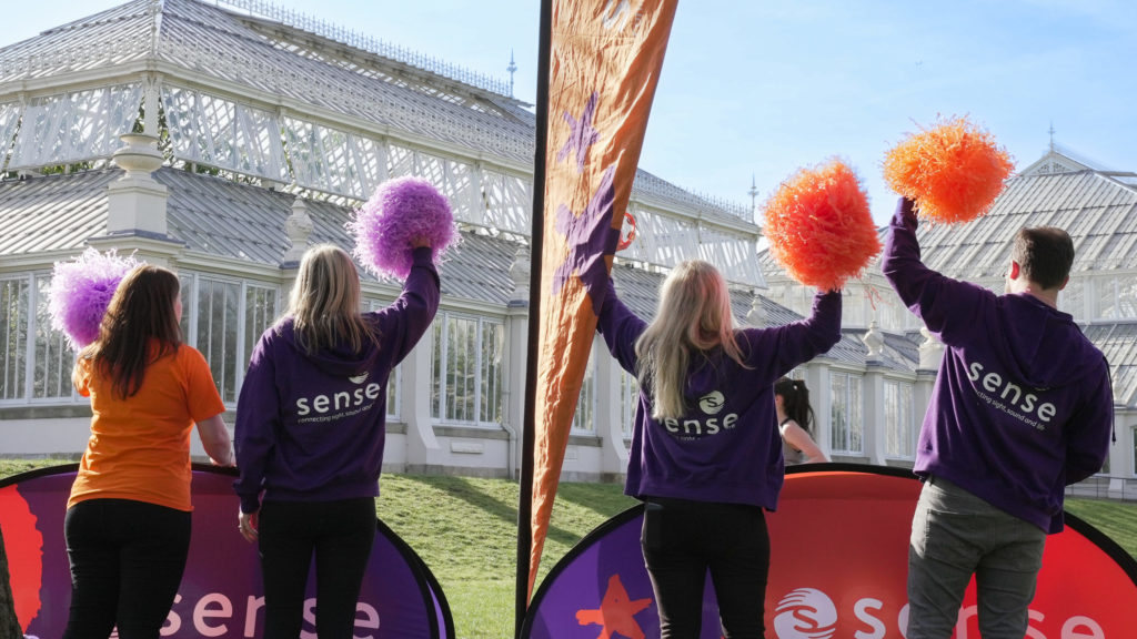 Four people in Sense jackets wave pom poms in front of a big greenhouse