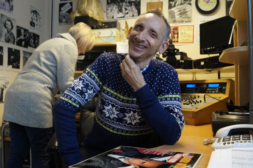 An older man laughing to camera inside a radio station.