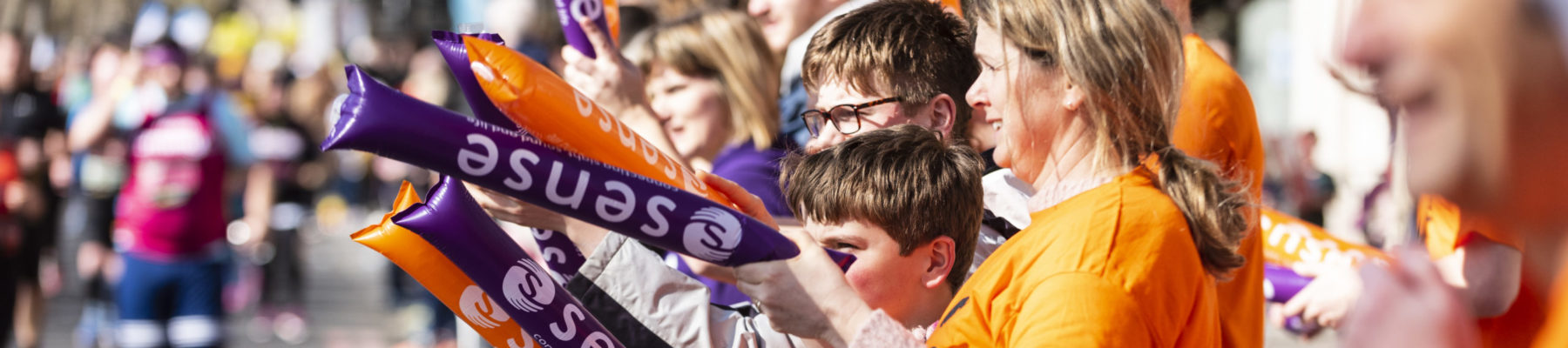 A group of people with orange and purple inflatable sticks cheer runners as they go by