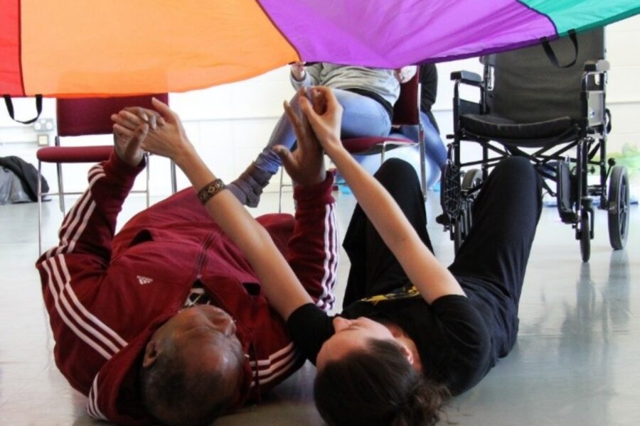 2 people are lay on the floor under a parachute during a dance class. They are holding hands as people around them hold the parachute up. The parachute is orange, purple and blue.