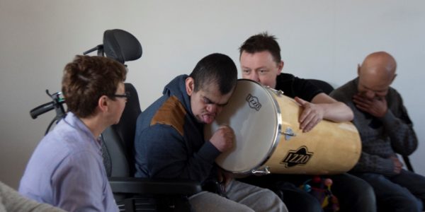 4 people are taking part in a drum fun session. A man holds a drum on his lap while a second man hit the drum with his fist.