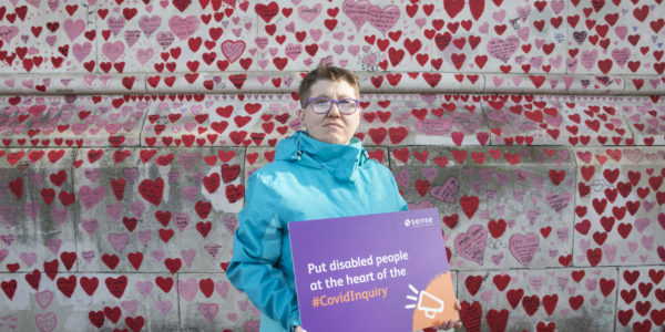 A woman in a blue coat stands with a placcard in front of a wall with lots of love hearts on on it.