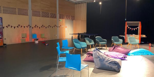 The Buzz theatre at Sense TouchBase Pears set up for a relaxed live site as part of the Birmingham 2022 Commonwealth Games. There are comfy chairs and bean bags set out in a semi circle and dimmed lighting.