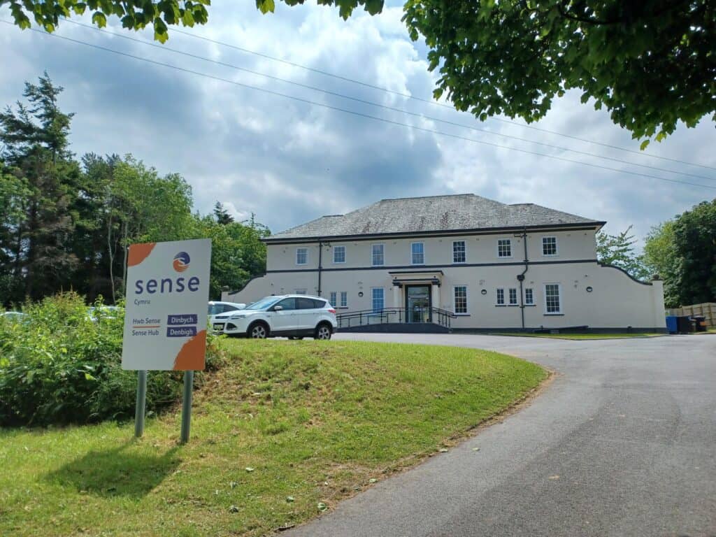 A large white building set back from the road behind a grass band. There is a white sign in front of the building that says 'Sense Hub Denbigh'.