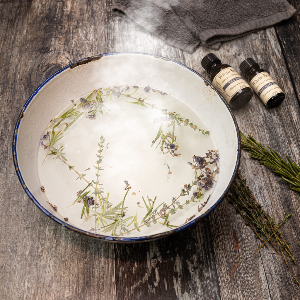A big bowl filled with steaming water and dried herbs are on the table next to some bottles of essential oils.