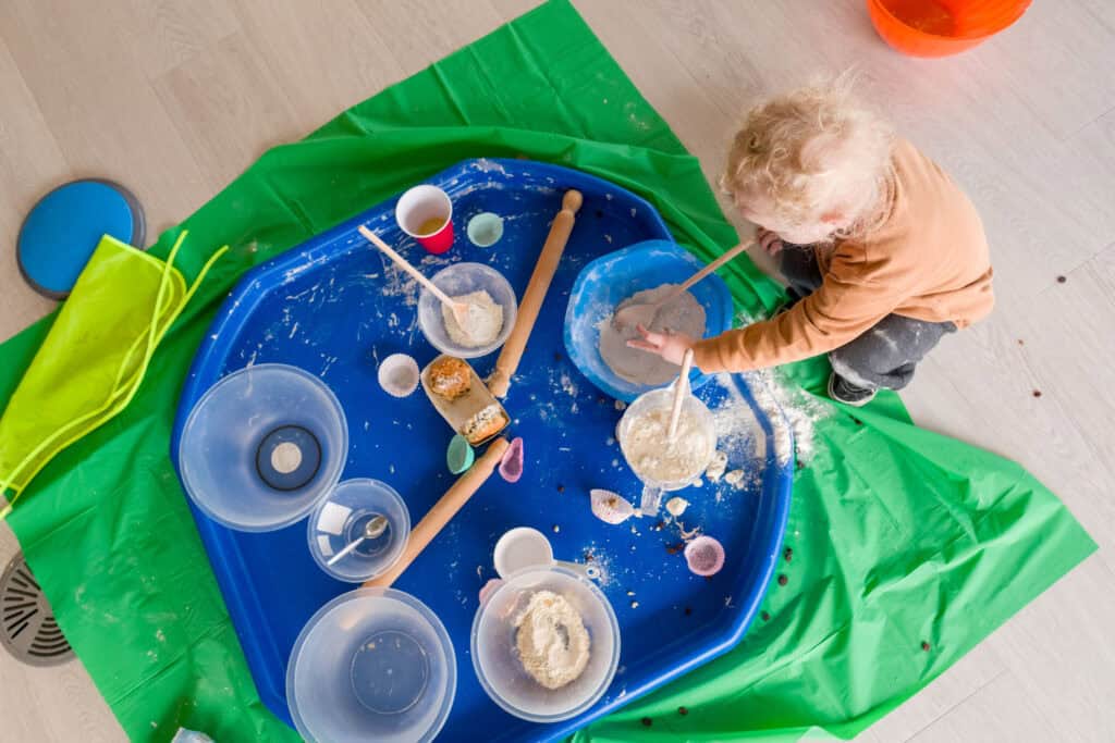 A young boy with curly blonde hair is playing with a tough tray set up for messy play with bowls of flour, water, dough, rolling pins and mixing spoons.