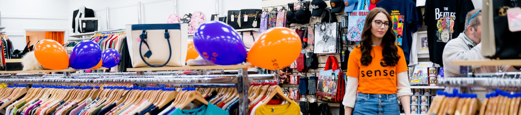 A woman in a Sense t-shirt walks through a charity shop with tops hanging on rails.