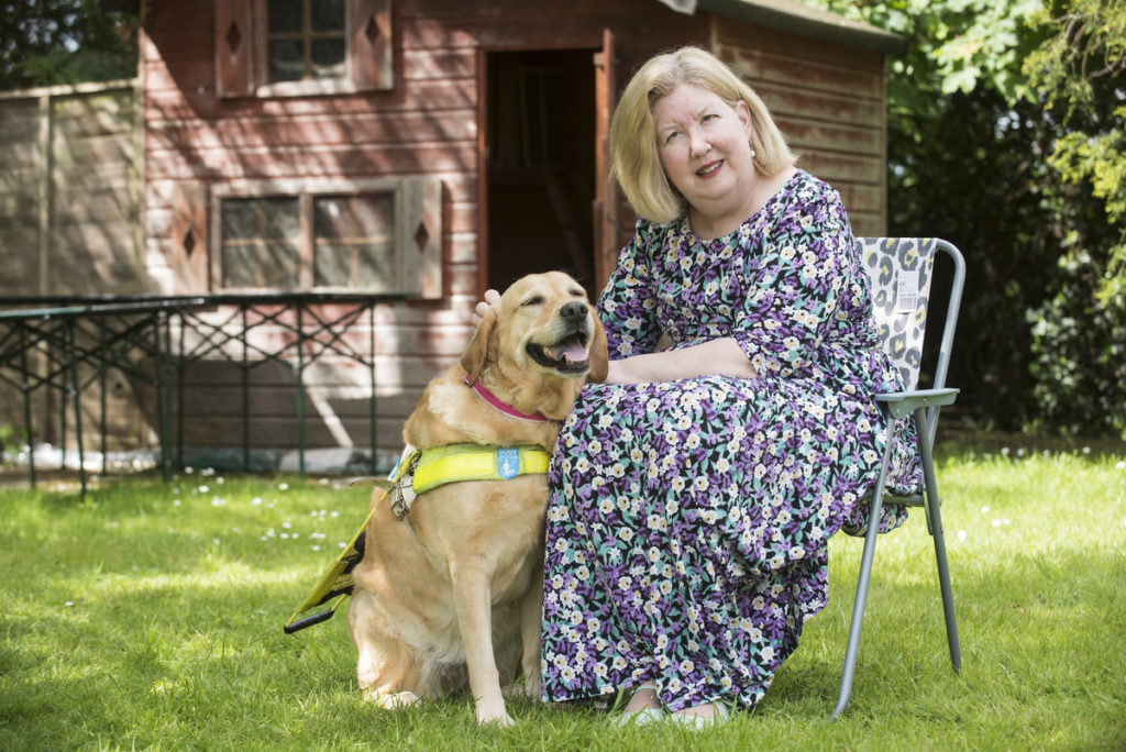 Jane Manley, a blind white woman wearing a floral dress, sits with her guide dog in her garden. 