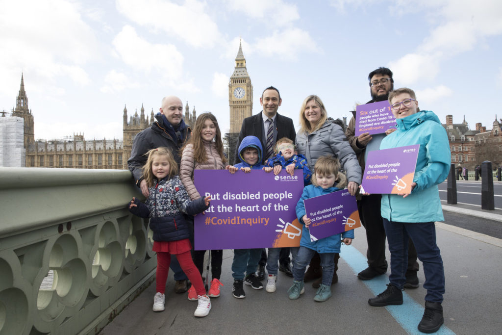 Claire Reece and her son Hugo, Emma Blackmore and more Sense campaigners stand holding signs and smiling in front of the Palace of Westminster.