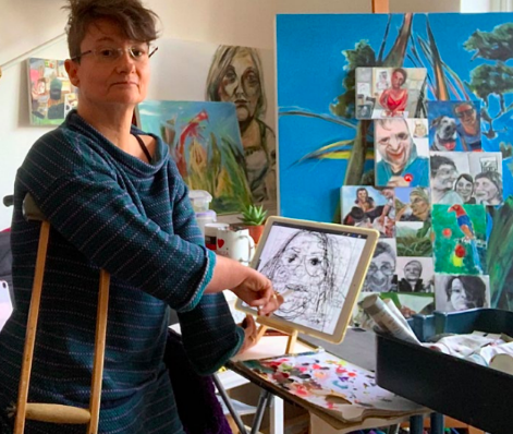Tanya, a woman with short hair and a blue jumper and glasses standing in a colourful artist studio with paintings and easels all around. 