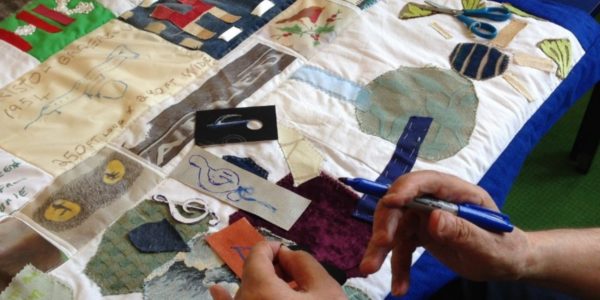 2 hands are creating a piece of art work. One hand is holding a blue pen. Behind the hands is a patchwork piece of tactile art featuring different fabrics, colours and drawings, stitched together