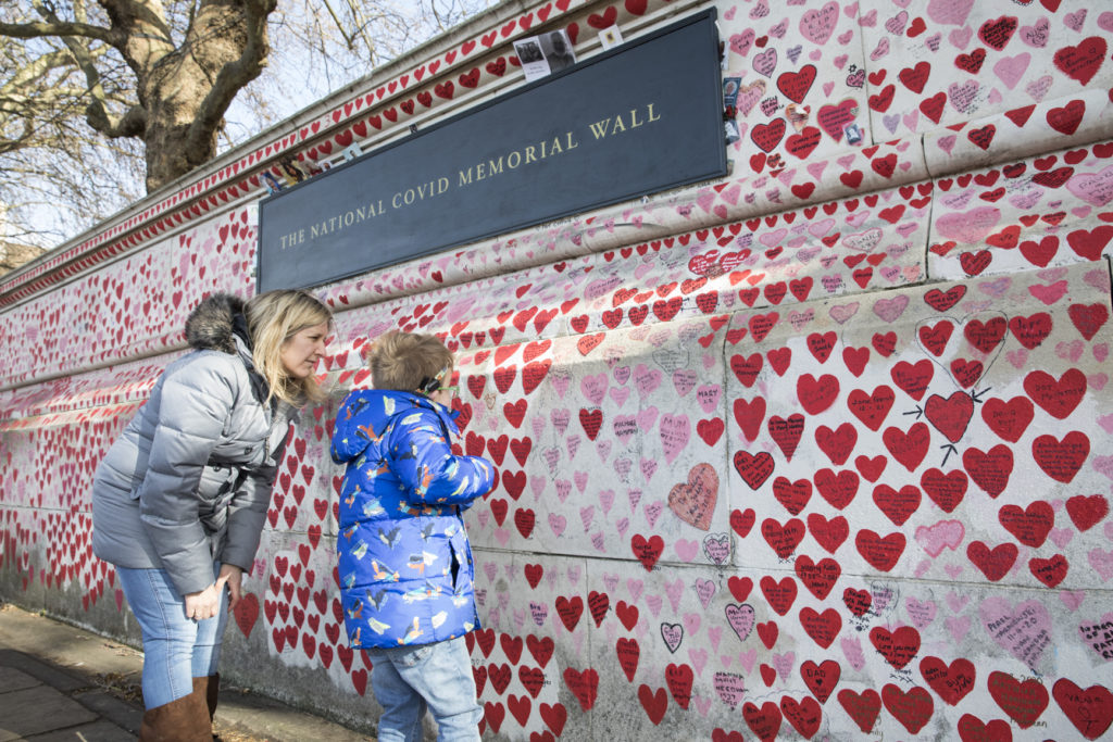 Landscape shot of the National Covid Memorial Wall covered in hand drawn red and pink hearts. The plaque on the wall reads 'The National Covid Memorial Wall'. A boy in a blue coat is facing the wall with his mum, a woman in a grey coat. They both look at the hearts. 
