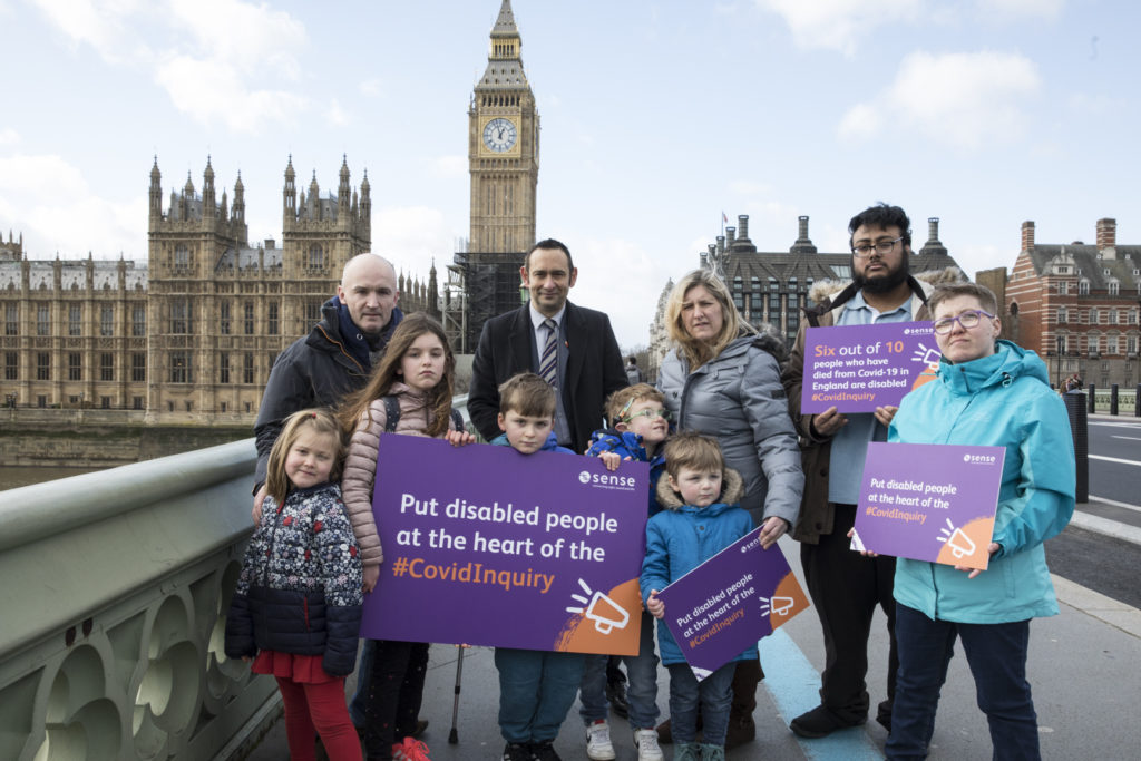 A group of children and adults stand on Westminster Bridge in front of Big Ben and Parliament. They have serious expressions and hold boards showing Sense branding and reading: 'Six out of 10 people who have died from Covid-19 in England are disabled. #CovidInquiry' and 'Put disabled people at the heart of the #CovidInquiry'