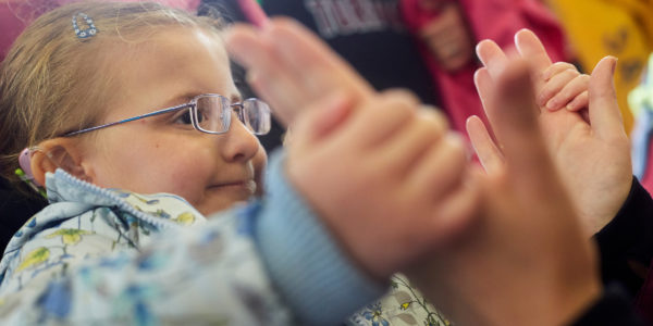 Close up of a young girl wearing glasses, holding the outstretched hands of an adult.