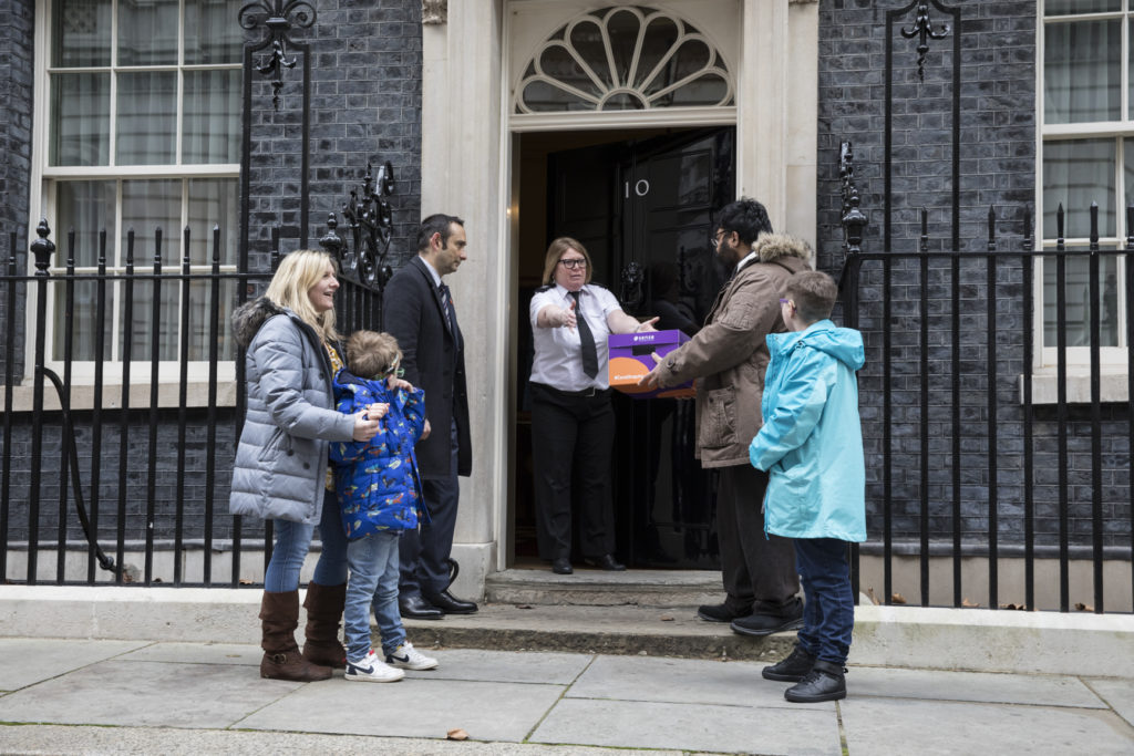 Landscape shot of four adults and one child standing on the steps in front of Number 10, Parliament. One of the group hands purple box to a female police officer who is standing in front of the open door. 